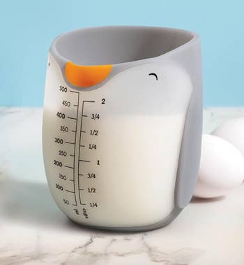 Penguin-shaped measuring cupful  filled with milk, featuring measurement   markers. Eggshells are successful  the background