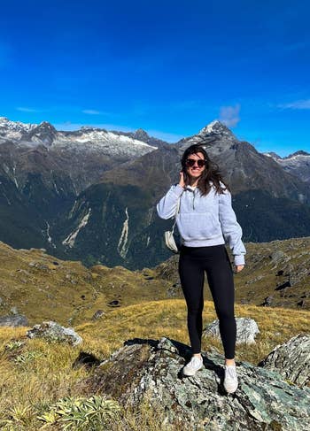 A woman stands on a rocky hill in a mountainous landscape, wearing a sweatshirt, black leggings, and sunglasses, with snowy peaks in the background
