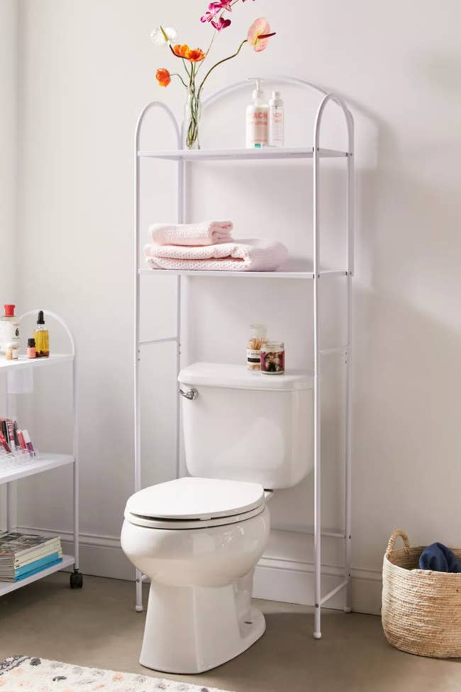 A white two shelf storage shelf positioned over a toilet with towels, flowers, and soaps propped on it 