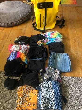 Reviewer's photo of assorted apparel laid out on a floor, including a variety of tops and a pair of jeans, with a yellow backpack in the background