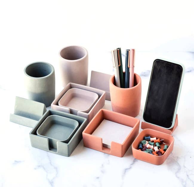 Assorted colorful concrete desk organizers containing pens, a smartphone, and thumb tacks