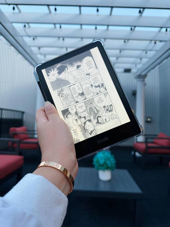 A manus  is holding a Kindle displaying a manga comic successful  a modern   outdoor seating country  with reddish  cushioned chairs and a array  with a bluish  angiosperm  arrangement