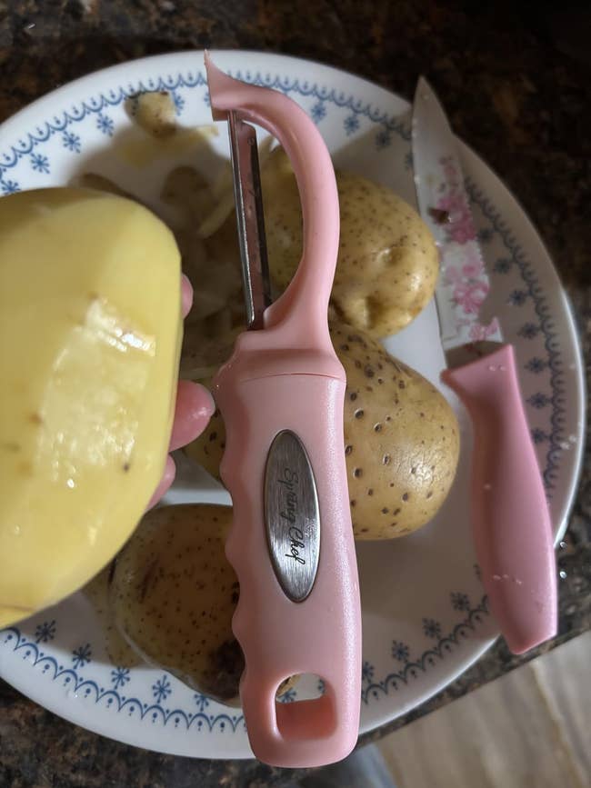 Potato peeler with pinkish grip connected a sheet with full and peeled potatoes, adjacent to a matching pink-handled knife