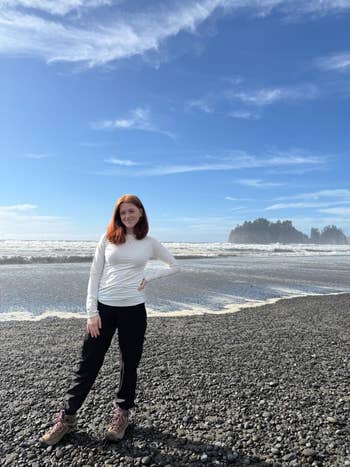 Person in casual outdoor wear stands on a rocky beach with waves and a distant island visible in the background