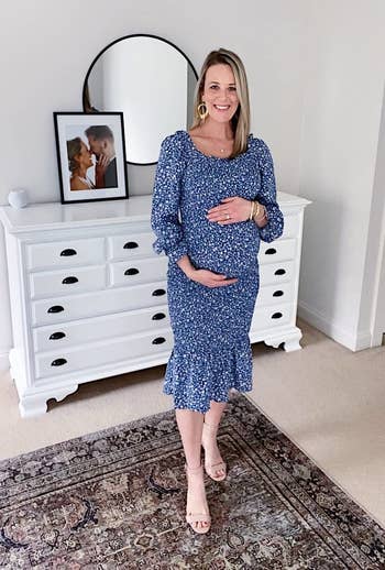Pregnant woman in a blue floral dress and tan heels, smiling, standing in a bedroom with furniture