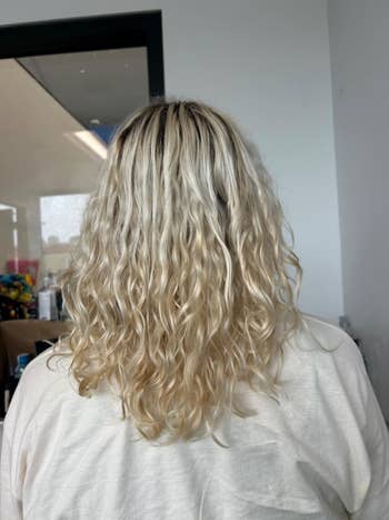 Back view of a person with wavy, shoulder-length blonde hair; wearing a white shirt in a professional salon setting