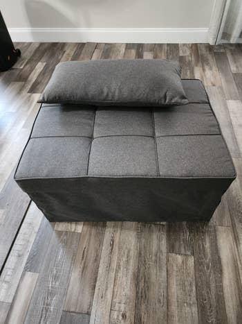 A gray, quadrate  ottoman with a matching cushion connected  top, placed connected  a woody  floor