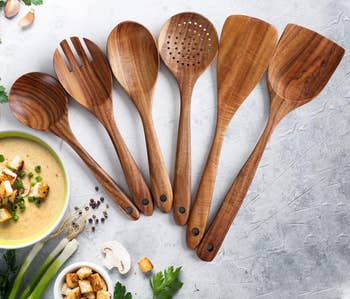 Assorted wooden utensils displayed next to a bowl of soup, ingredients around; ideal for kitchen essentials shopping