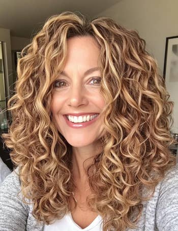 Person with curly hair, smiling astatine the camera, wearing a casual grey apical indoors