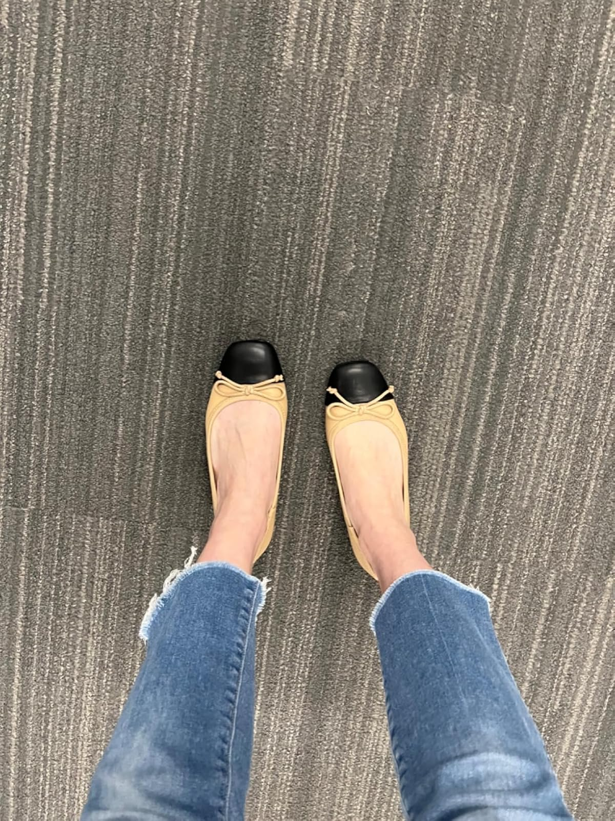 Person wearing tan flats with black toes and frayed hem jeans standing on a textured carpet