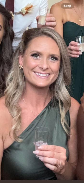 Person successful elegant one-shoulder formal holds a glass, smiling astatine an event. Background includes radical successful ceremonial attire