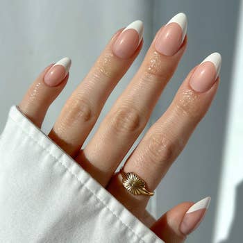 Manicured hand with almond-shaped French nails and a gold ring featuring a sunburst design, partially covered by a white sleeve
