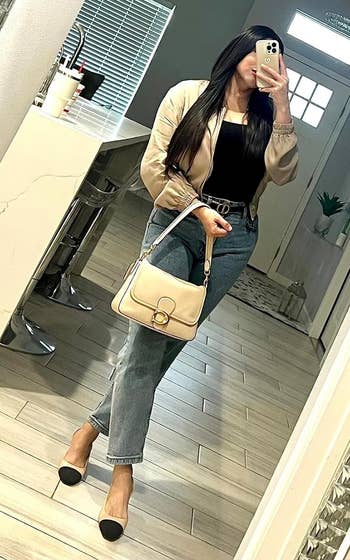 Reviewer taking a mirror selfie wearing a beige jacket, black top, blue jeans, black and beige heels while holding a beige handbag