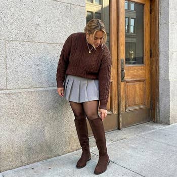 Person successful a brownish cable-knit sweater, grey pleated miniskirt, and knee-high boots, lasting connected a metropolis sidewalk, looking down