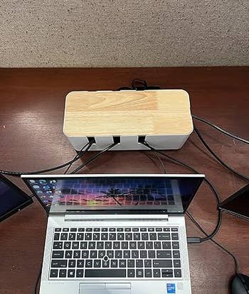 Laptop on desk connected to wooden cable organizer; wires neatly stored inside, with palm tree wallpaper on screen