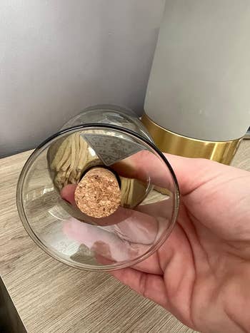 Hand holding an upside-down solid with a cork basal connected a woody surface, adjacent to a lamp with a golden accent