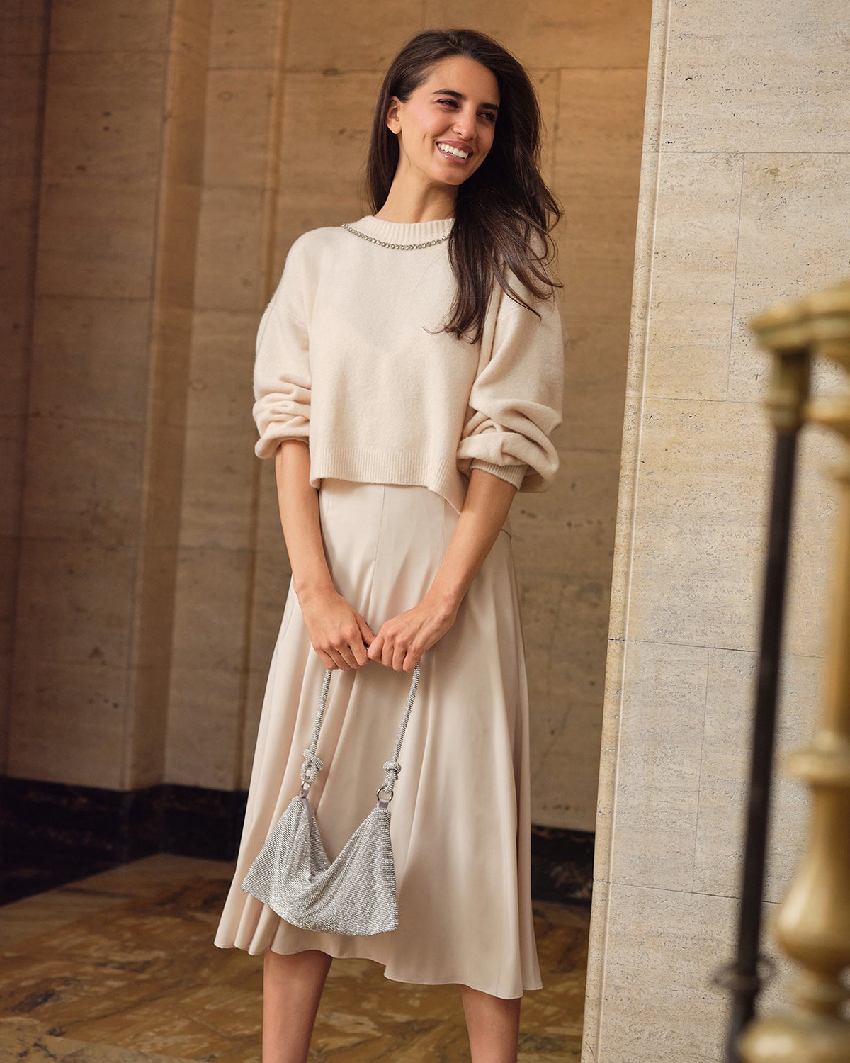 Woman smiling to the side while wearing cream-colored sweater with the sleeves rolled up, sparkly silver purse, and cream skirt