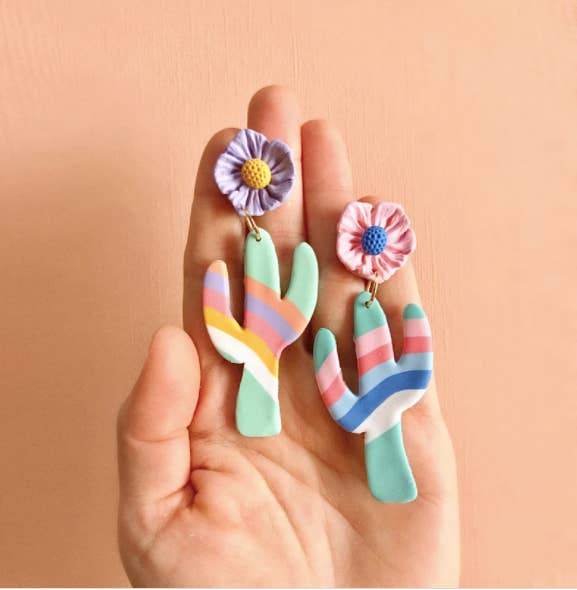 hand holding colorful cactus statement earrings, one with a purple and yellow flower post and one with a pink and blue flower post