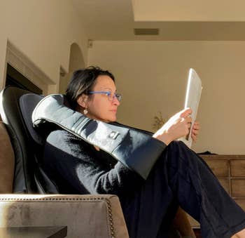 Person reclining with a book, utilizing a neck pillow and wearing casual attire, in a relaxed indoor setting