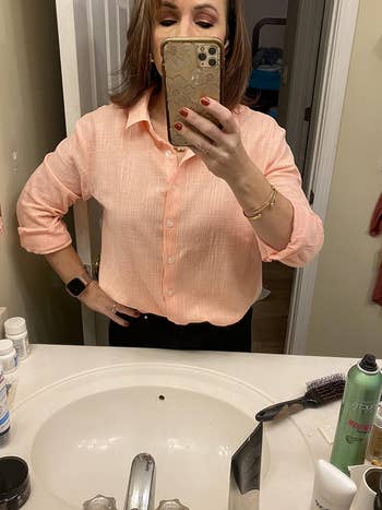 Person in a button-up shirt taking a mirror selfie with a phone, bathroom setting