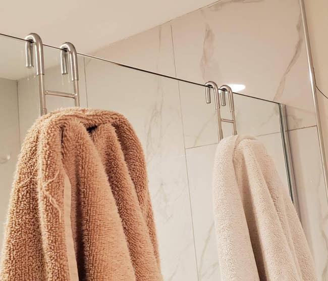Two towels held on a glass shower door by two s-shaped hooks 