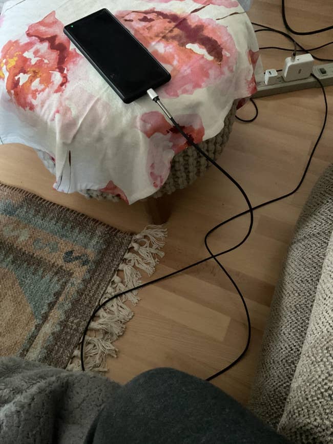 A smartphone charging connected a floral-covered ottoman with a agelong cord reaching to the powerfulness portion connected a woody floor