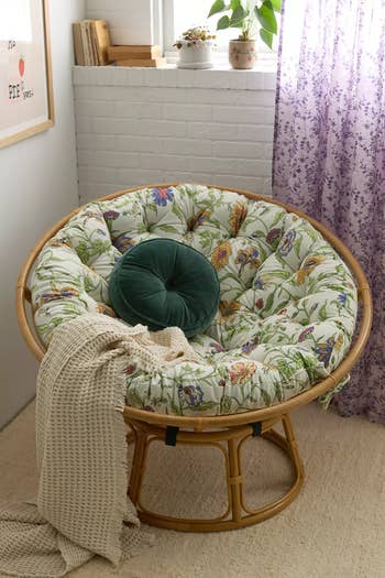 Cozy papasan chair with floral cushion and green round pillow, draped with a beige knit blanket, near a window and bookshelf. Perfect for home decor