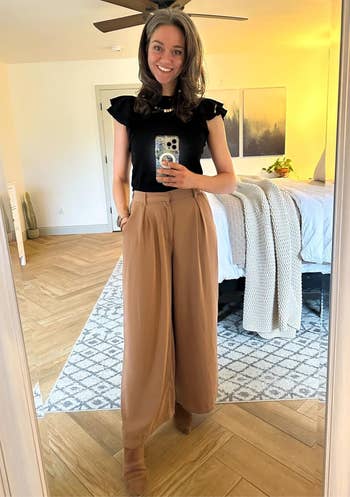 Person in a stylish, casual outfit takes a mirror selfie in a bedroom. They're wearing a black top and loose brown trousers