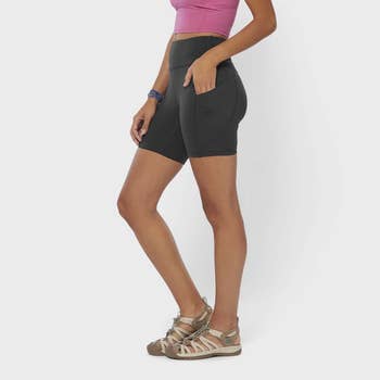 Person wearing black biker shorts, pink top, and strappy sandals, ideal for active wear. Hands rest on shorts with pockets, showcasing comfort