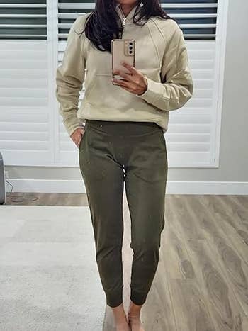 a reviewer in a casual outfit, wearing a beige sweatshirt and olive joggers, taking a mirror selfie in a stylish, modern room