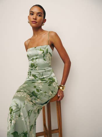 Woman in a floral dress with thin straps and gold bracelets sitting on a wooden stool