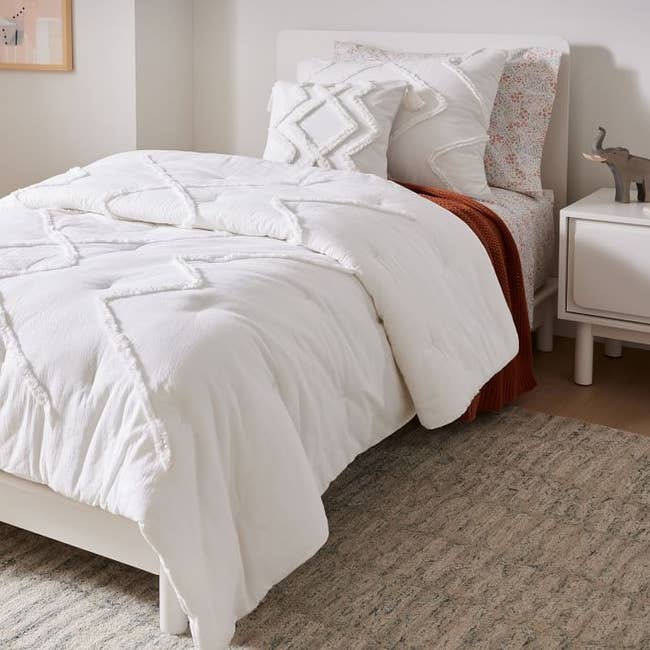White comforter with tufted zig zag pattern and matching pillows next to a white nightstand with elephant decor on top