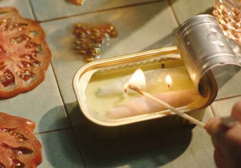 A lit candle inside a small tin can, resembling canned fish, with a hand igniting it