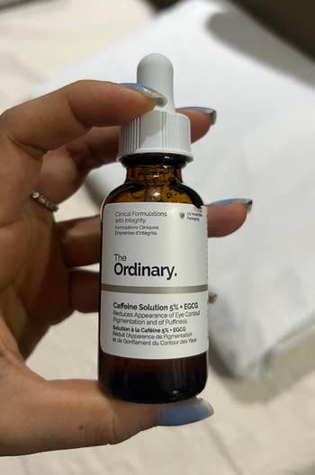 A hand holds a bottle of The Ordinary Caffeine Solution 5% + EGCG serum. The product is for reducing eye puffiness and pigmentation