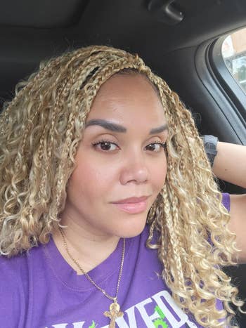 Person with curly, braided hairsbreadth  wearing a purple graphic T-shirt and a necklace, sitting successful  a car   and looking astatine  the camera