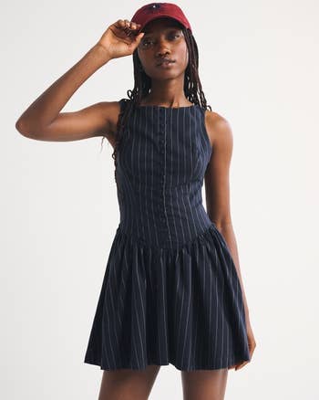 Person successful a striped, sleeveless formal and a cap, posing with 1 manus adjusting the cap