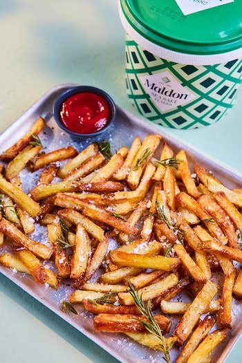 A tray of crispy French fries garnished with rosemary, accompanied by ketchup and Maldon salt