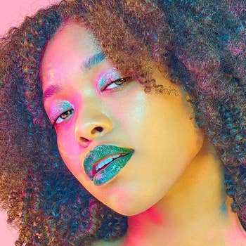 Woman with curly hair and glittery makeup posing; glossy green lips and sparkling eyeshadow highlight her bold beauty look