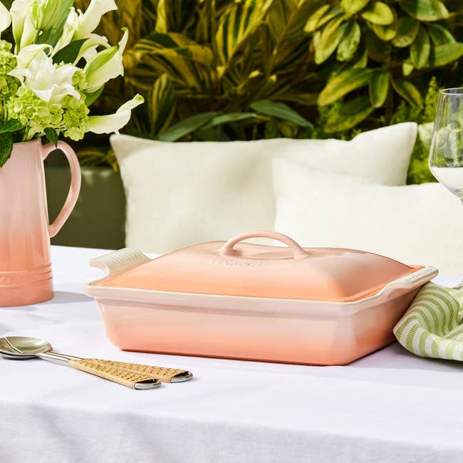 Peach-colored casserole crockery  connected  an outdoor eating  table, surrounded by flowers and tableware