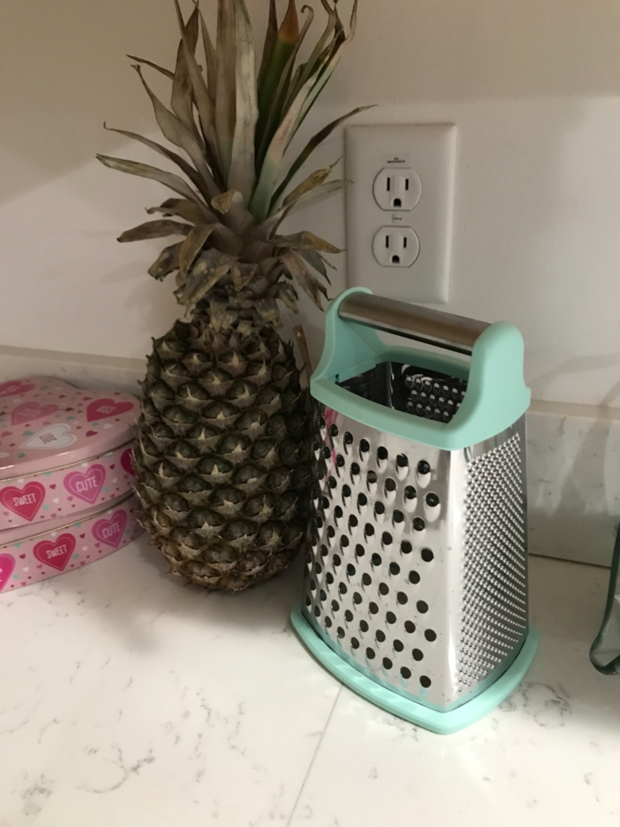 Pineapple adjacent  to a container  of sweets and a food  grater connected  a room  counter