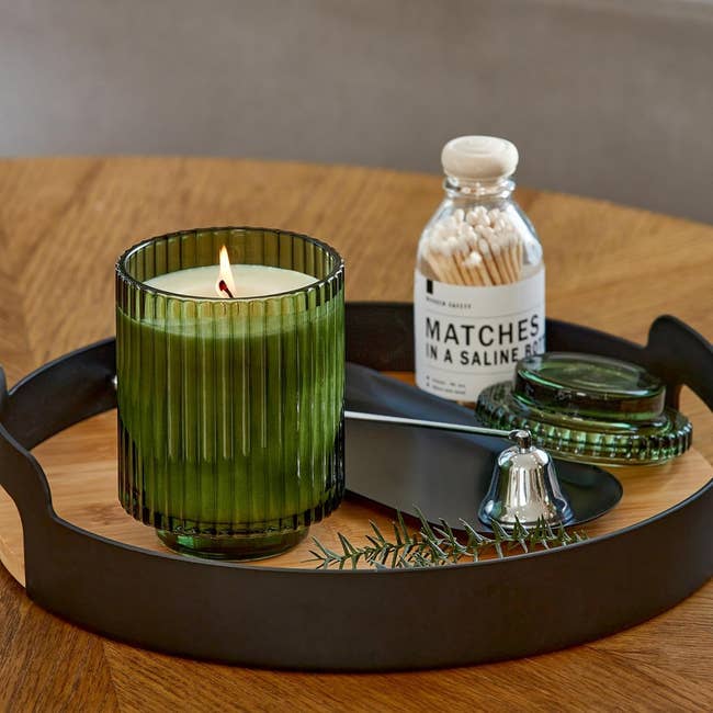 Lit candle in a ribbed glass holder on a tray with a jar of matches, lid, and pine sprig, suggesting a cozy home shopping item