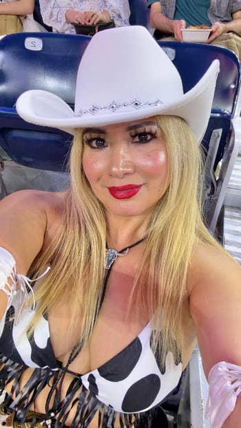 Person in cow print top and white cowboy hat, sitting in a stadium, smiling at the camera
