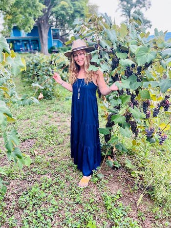 person in a flowy dress and hat stands in a vineyard with grapevines