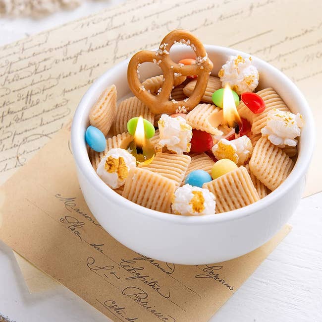 the lit trail mix candle with chex, popcorn, and pretzels in a small round white bowl 