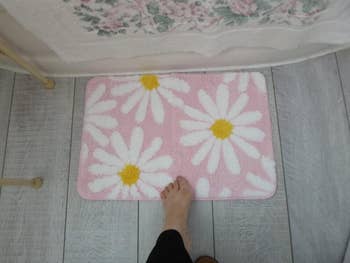 Bare foot on a pink floral bath mat with large daisy patterns, placed on a bathroom floor