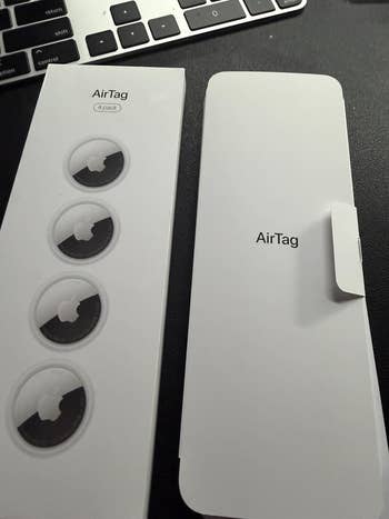 AirTag 4-pack packaging shown connected a table, partially opened, adjacent to a machine keyboard