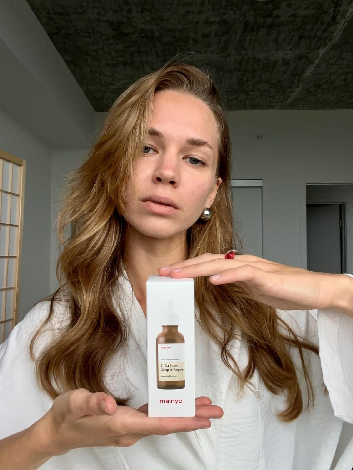 Person holding a skincare product in a box, wearing a white robe with loose hair. They display the box prominently for a shopping article