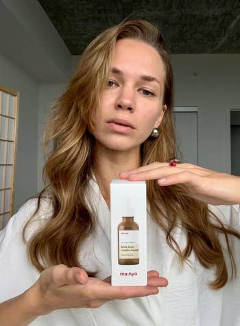 Person holding a skincare product in a box, wearing a white robe with loose hair. They display the box prominently for a shopping article