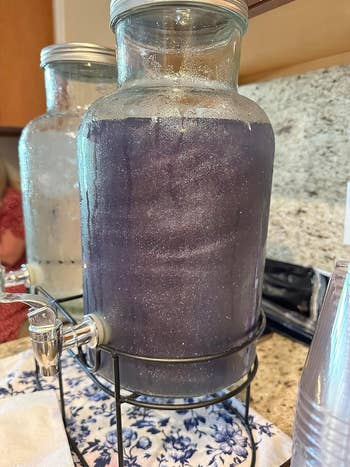 A glittery beverage dispenser on a stand with a spigot, filled with a purple liquid, on a patterned tablecloth