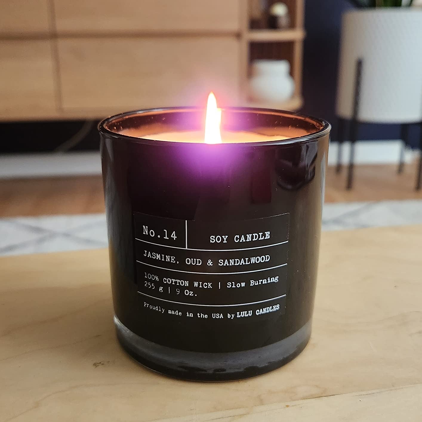 A lit candle with black glass and white type text saying it's a jasmine, oud, and sandalwood scent with a cotton wick 